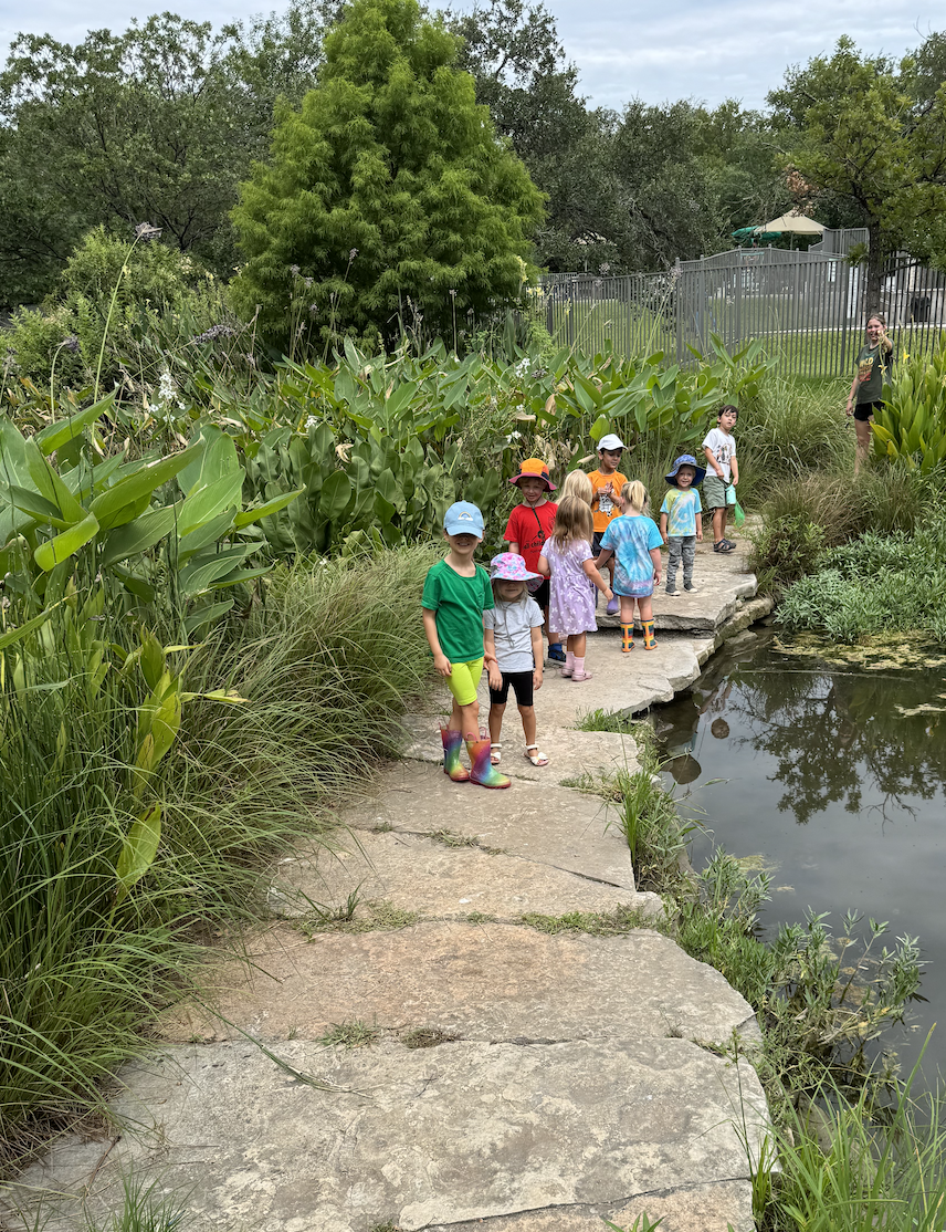 Children walking single-file along a stone path beside a pond with their teacher